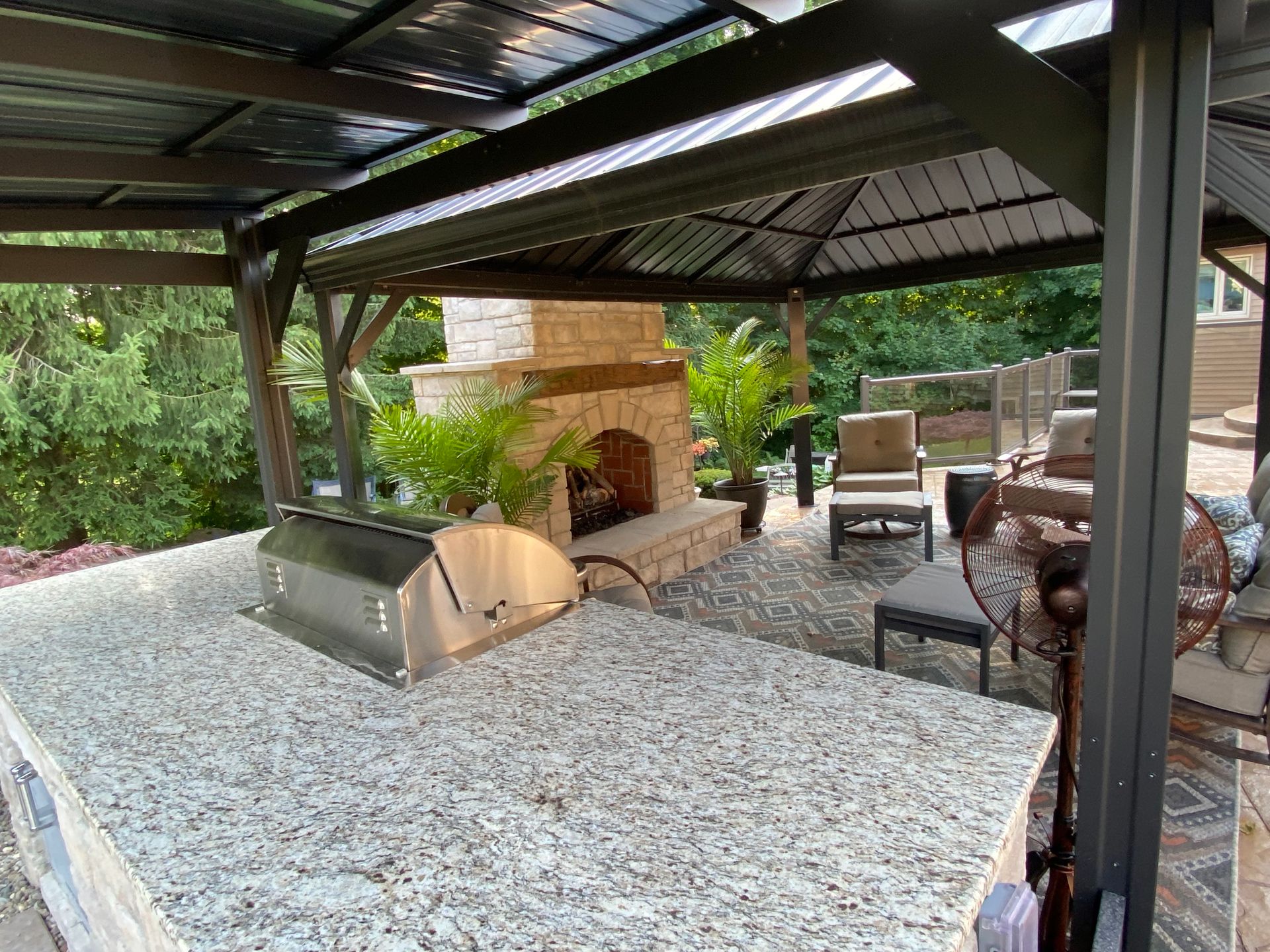Covered patio with stone fireplace, gravel floor, and outdoor seating surrounded by greenery