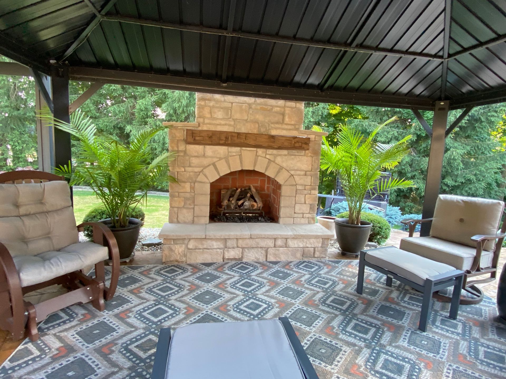 Covered patio with stone fireplace, cushioned chairs, patterned rug, and lush tropical plants.