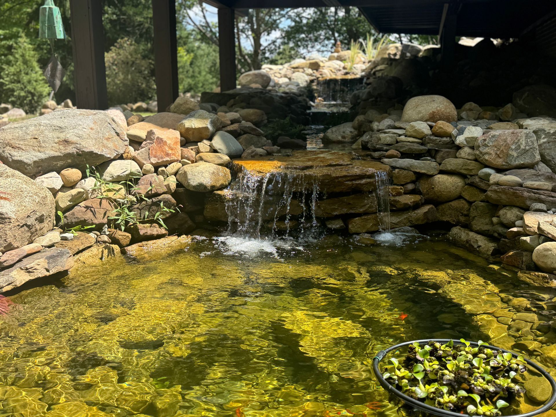Rock waterfall flowing into a clear pond with rocks and lily pads in a shaded garden pond.