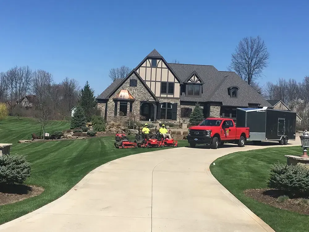 Large house with a curved driveway, red truck, and lawn equipment in a landscaped yard under a clear blue sky.