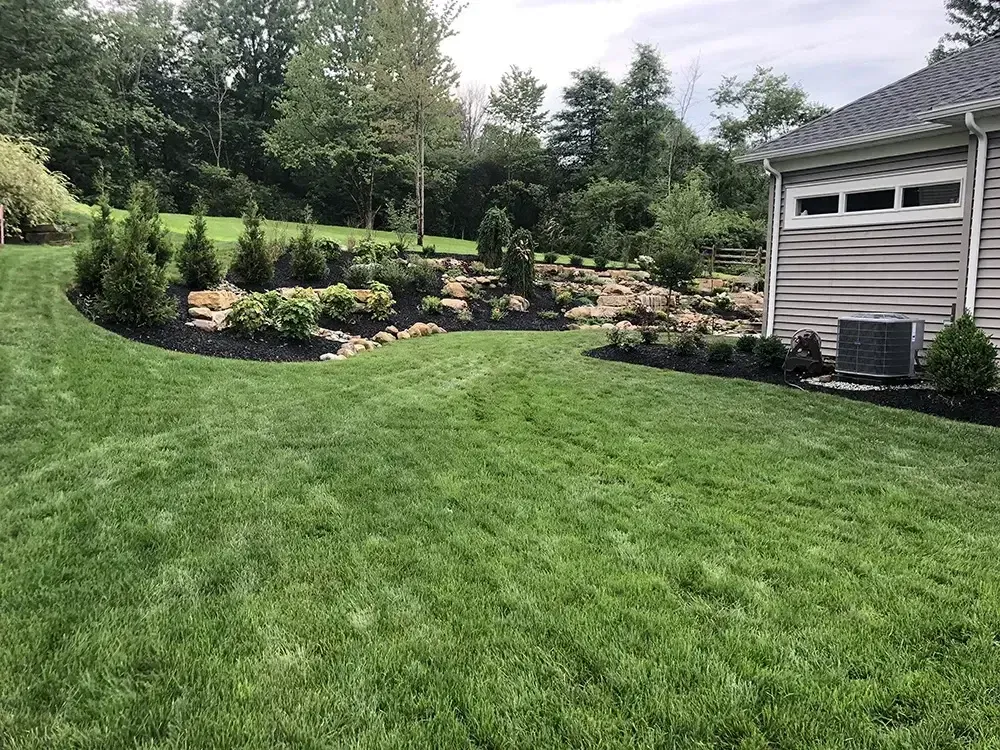 Lush green lawn with landscaped flower beds and trees beside a light-gray house.
