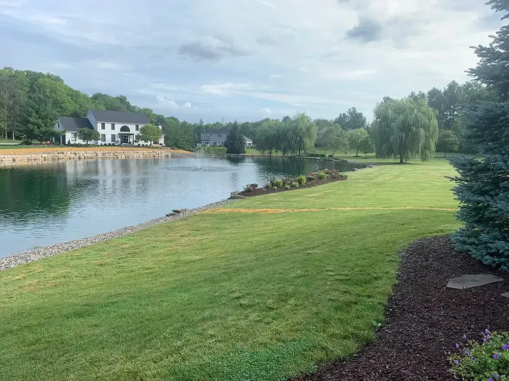 Quiet lakeside park with green lawn, trees, and houses across the water under a cloudy sky.