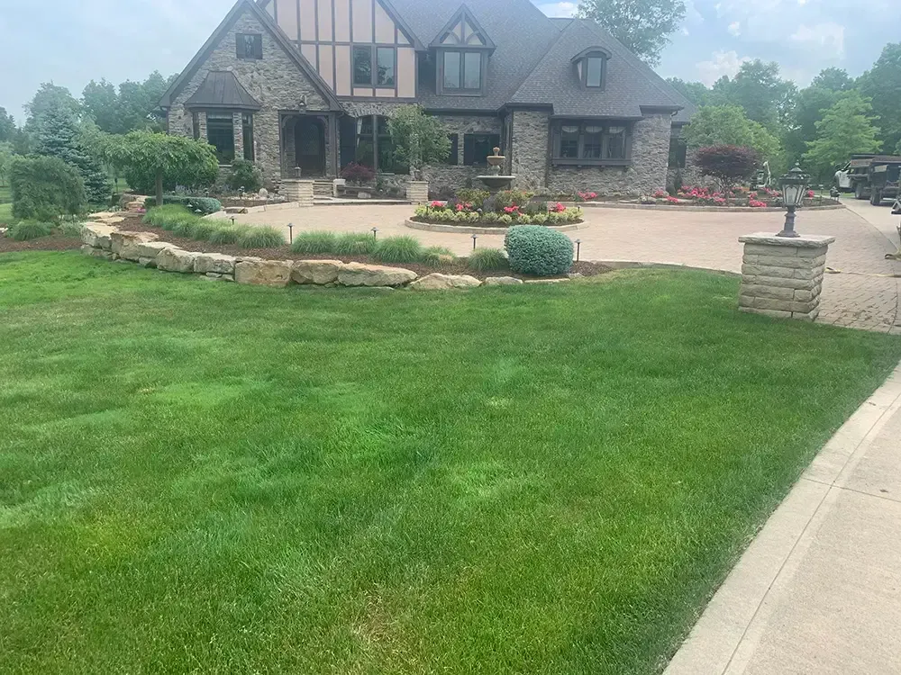 Large stone house with a manicured lawn, flower beds, and a curved driveway under a cloudy sky.