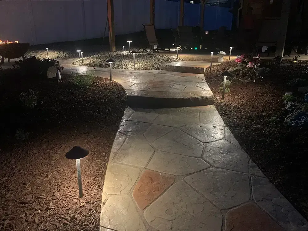 Nighttime garden path with stone pavers, landscape lights, and shrubs leading toward a lit patio area.