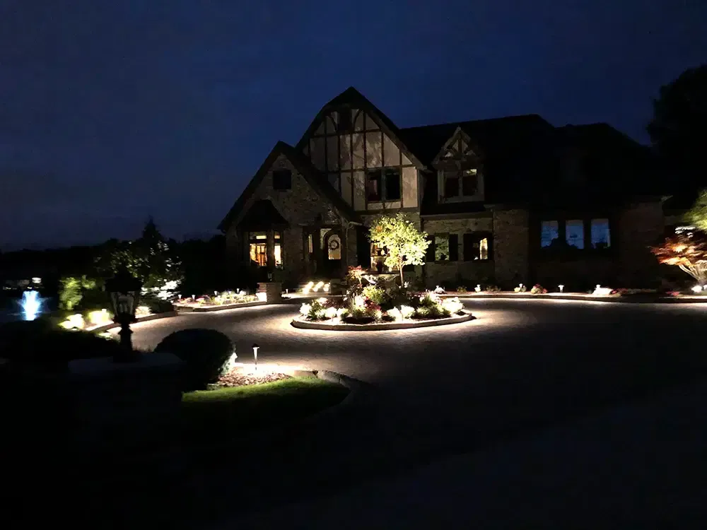 Nighttime view of a large house with illuminated landscaping and a lit driveway entrance.