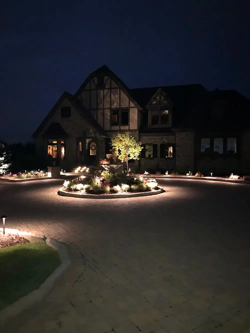 Illuminated Tudor-style house at night with a lit driveway and landscaped island garden.