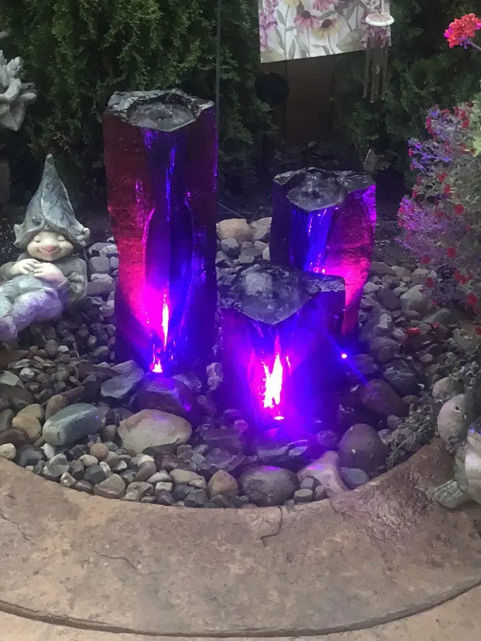 Three purple-lit water features in a rock garden, surrounded by pebbles and plants.