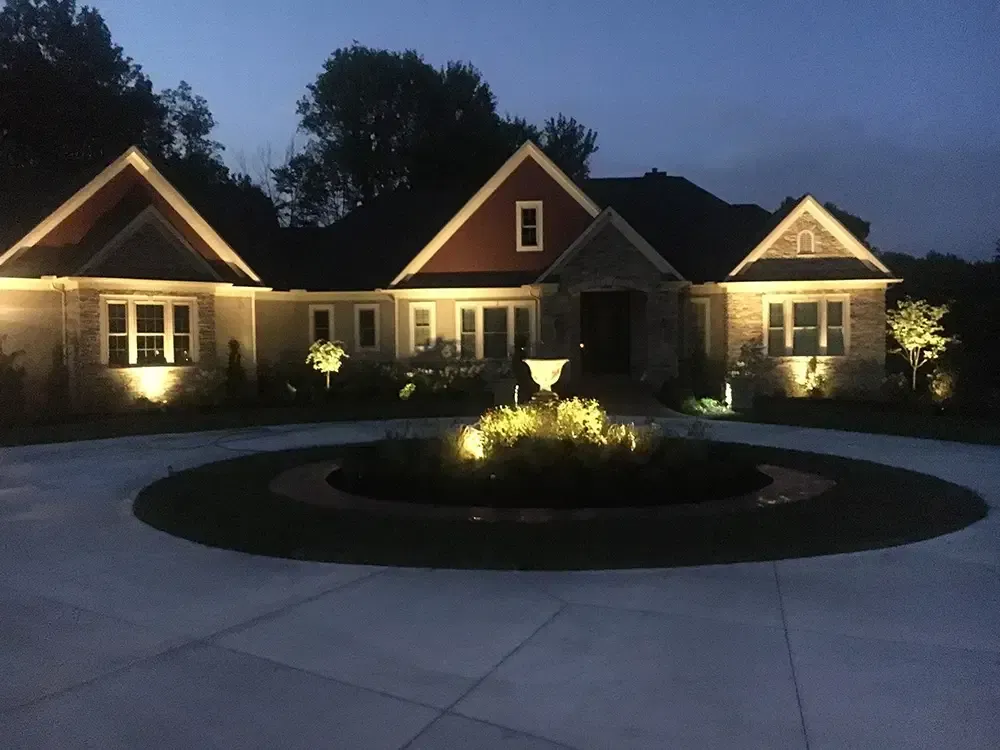 Illuminated house at dusk with lit windows, gabled roofs, and a circular driveway fountain centerpiece.