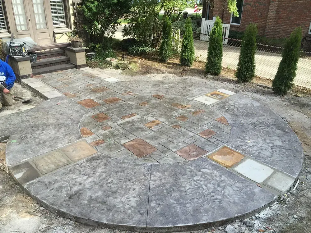 Circular stone patio with mixed pavers in a backyard, beside steps, shrubs, and a brick house.