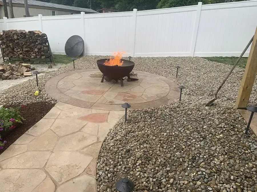 Backyard fire pit on a circular stone patio, surrounded by gravel and a white fence.