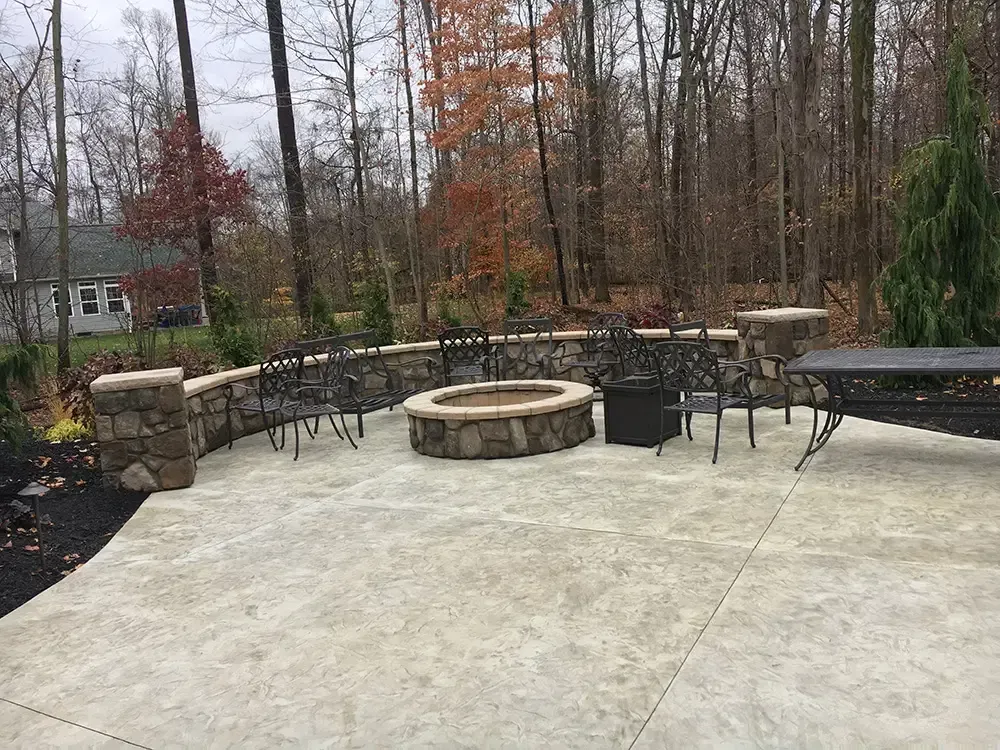 Stone patio with fire pit, outdoor seating, and wooded backyard in late fall.