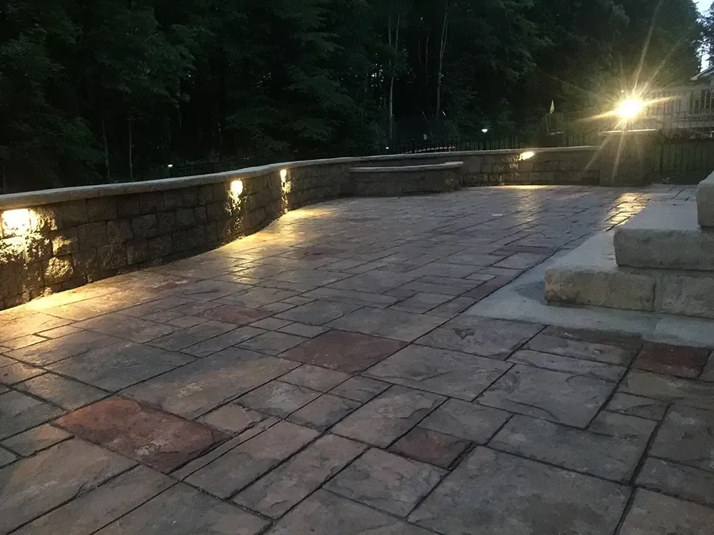 Stone-paved terrace at dusk with low wall lights and trees in the background.