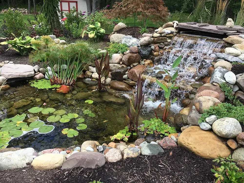 Garden pond with lily pads and a small waterfall over stacked rocks, surrounded by flowers and greenery.