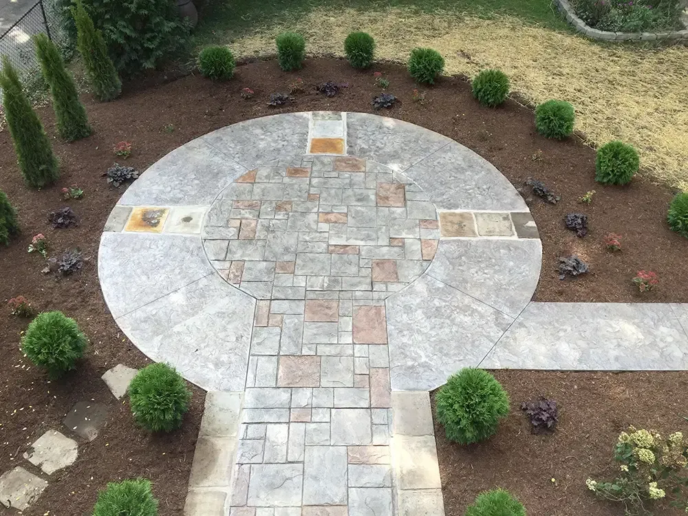 Circular stone patio with a cross-shaped walkway in a landscaped garden with shrubs and mulch.