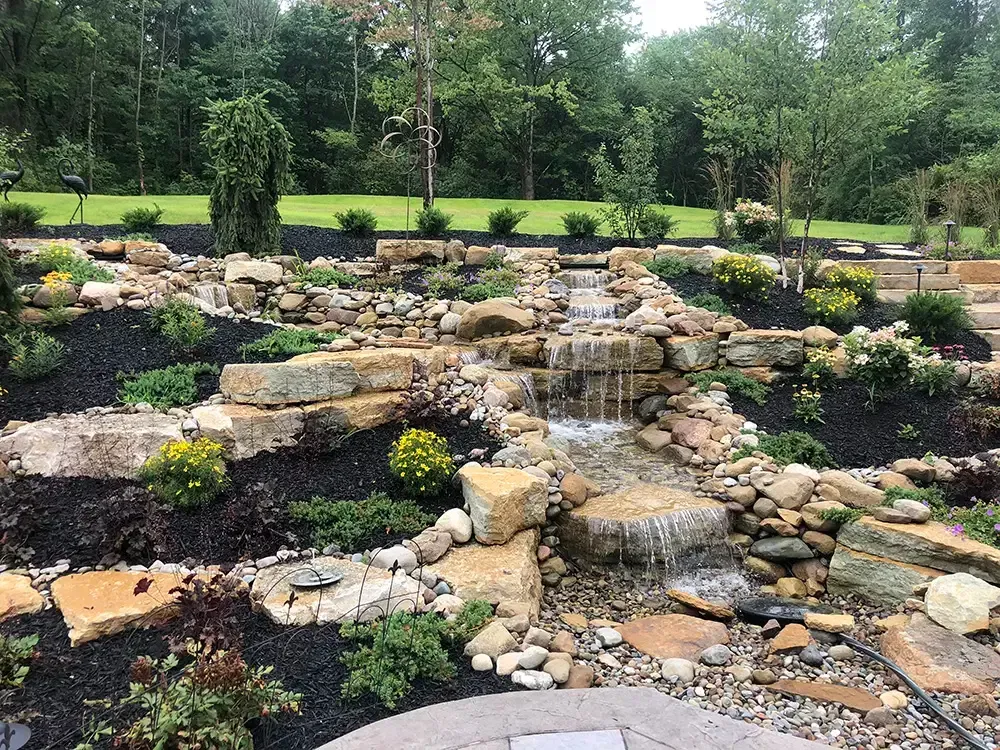 Tiered stone waterfall in a landscaped garden with rocks, greenery, and trees.