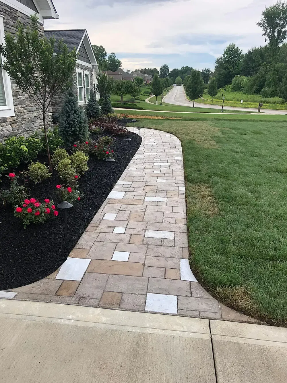 Brick walkway with white stepping stones curving past a house and flower beds to a green lawn and trees.
