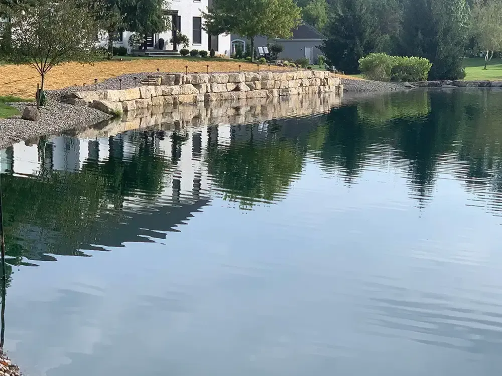 Calm lake reflecting trees and a stone embankment beside a white house on a sunny day.
