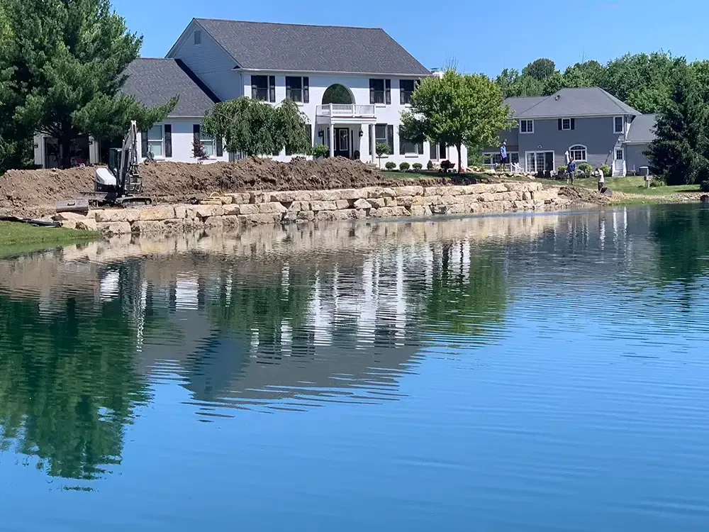 Waterfront house with white columns reflected in a calm lake, beside a rocky shore and green trees.