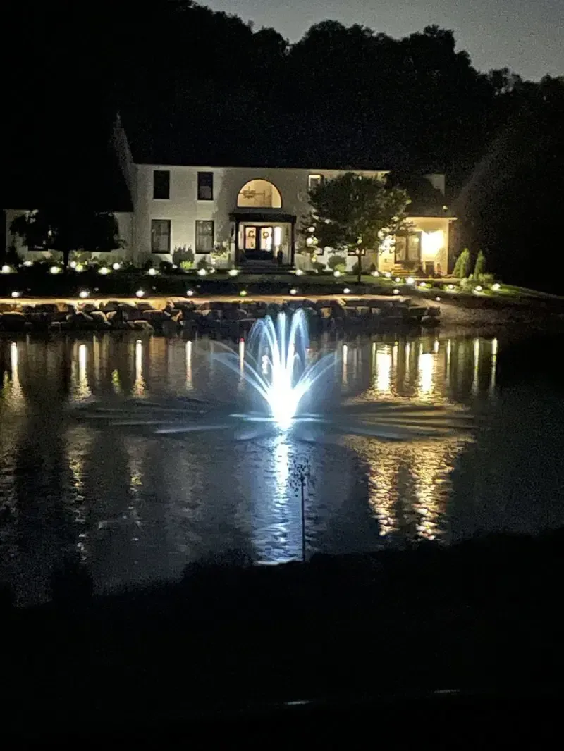 Nighttime view of a lit house and fountain reflecting on a pond.