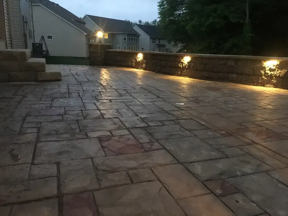 Stone patio at dusk with warm wall lights and houses in the background.