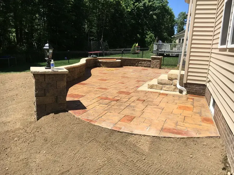 Patio with tan pavers, stone retaining wall, and sandy yard beside a house.