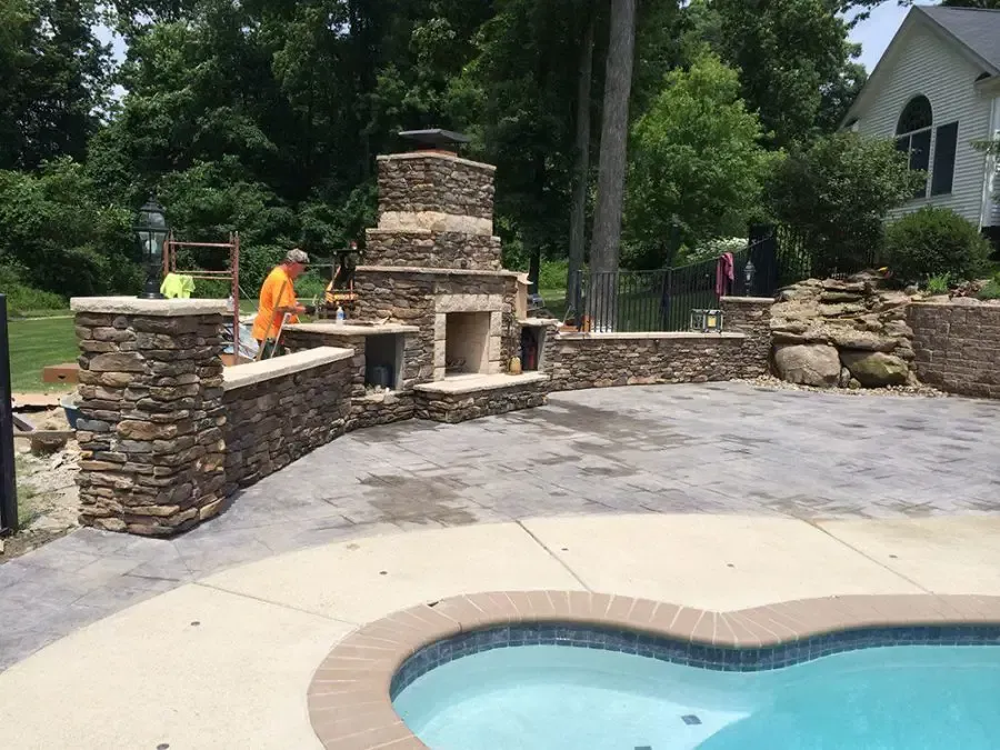 Stone outdoor fireplace and seating area beside a blue swimming pool, with workers nearby.