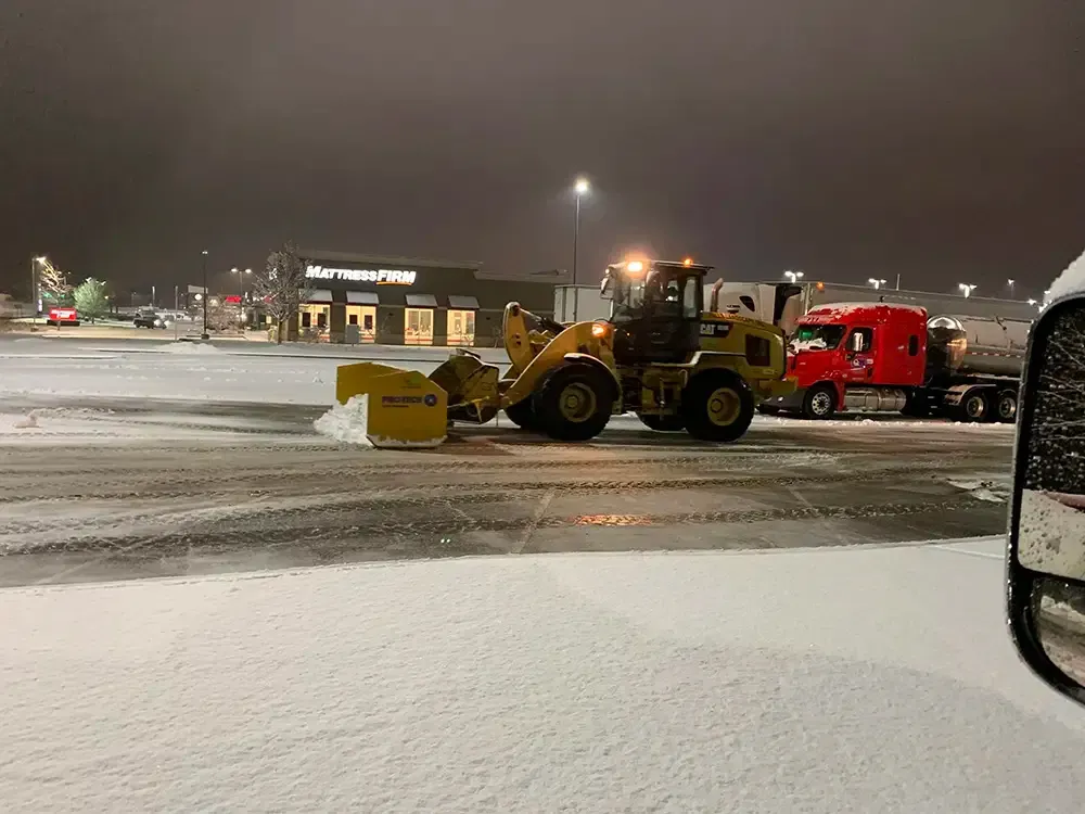 Snowplow clearing a snowy parking lot at night beside a red truck and lit storefronts.