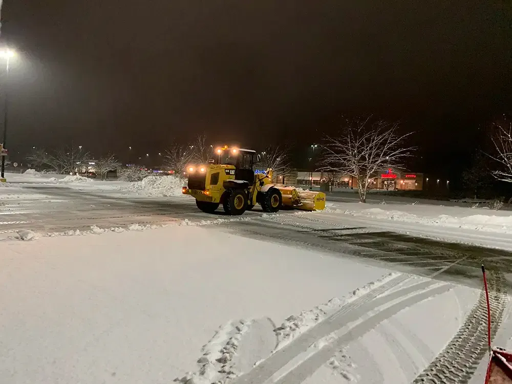 Yellow snowplow clearing a snowy parking lot at night under streetlights.