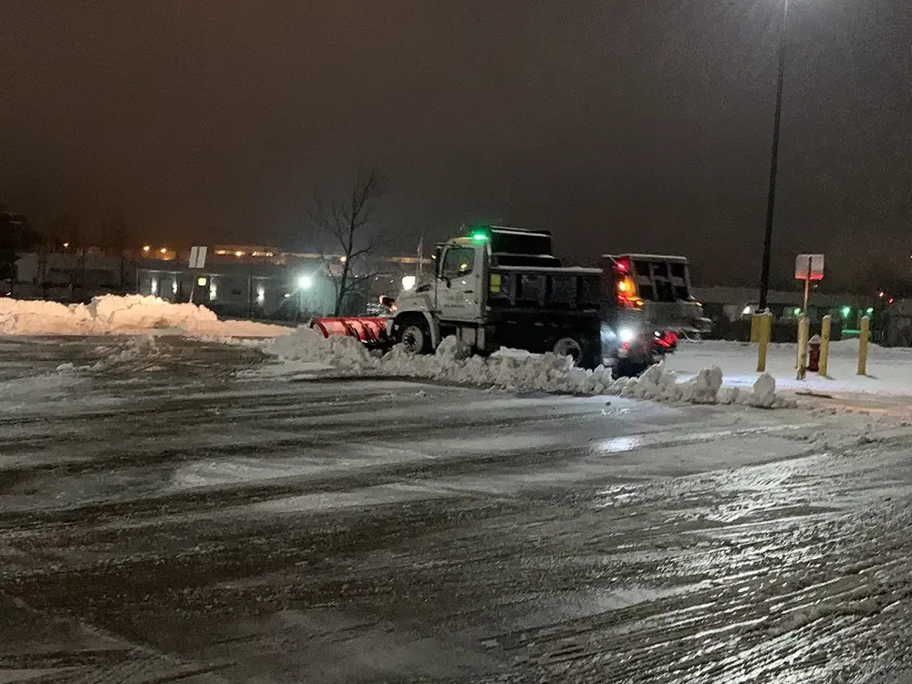 Tow truck clearing snow in a wet parking lot at night, with streetlights and parked vehicles nearby.