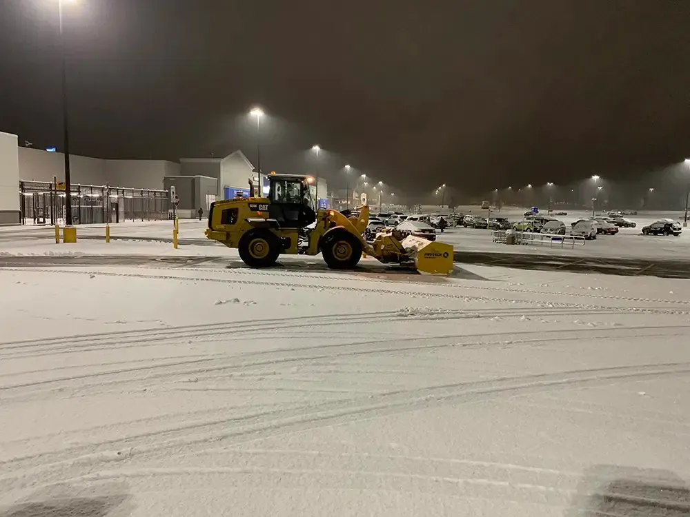 Yellow snowplow clearing a snowy parking lot at night under streetlights.