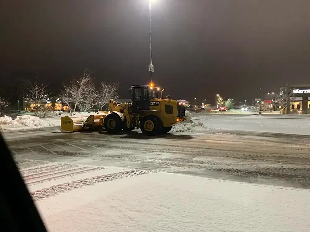 Yellow snowplow clearing a snowy parking lot at night under bright streetlights.