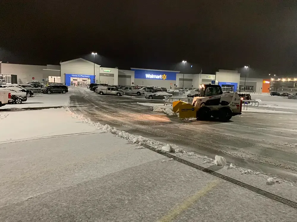 Snow-covered parking lot at night with a yellow snowplow clearing a curved path near a store.