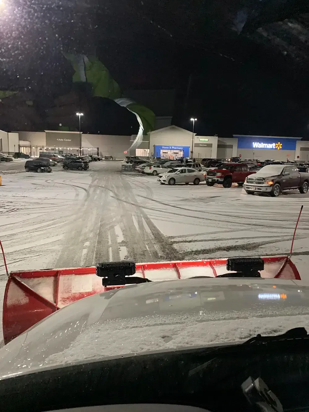 Snowy parking lot at night, viewed from inside a snowplow with red blade in the foreground.