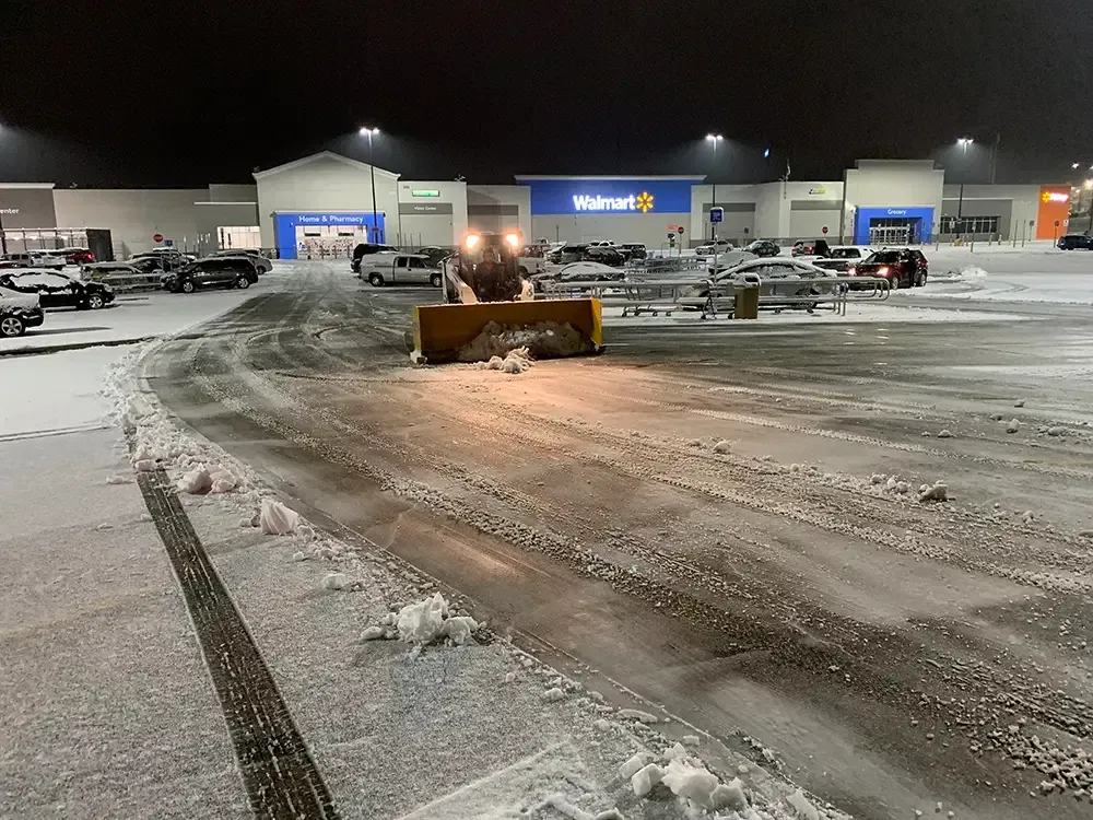 Snowplow clearing a snowy Walmart parking lot at night with store lights in the background.