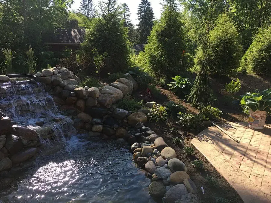 Rock garden waterfall beside a stone patio, surrounded by trees and sunlight.