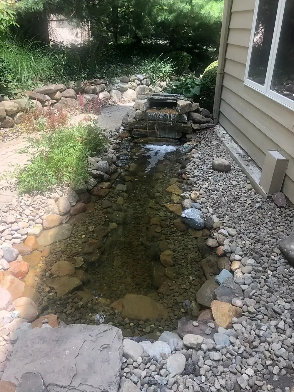 Rock-lined garden pond or dry creek bed with shallow water beside a house and shrubs.