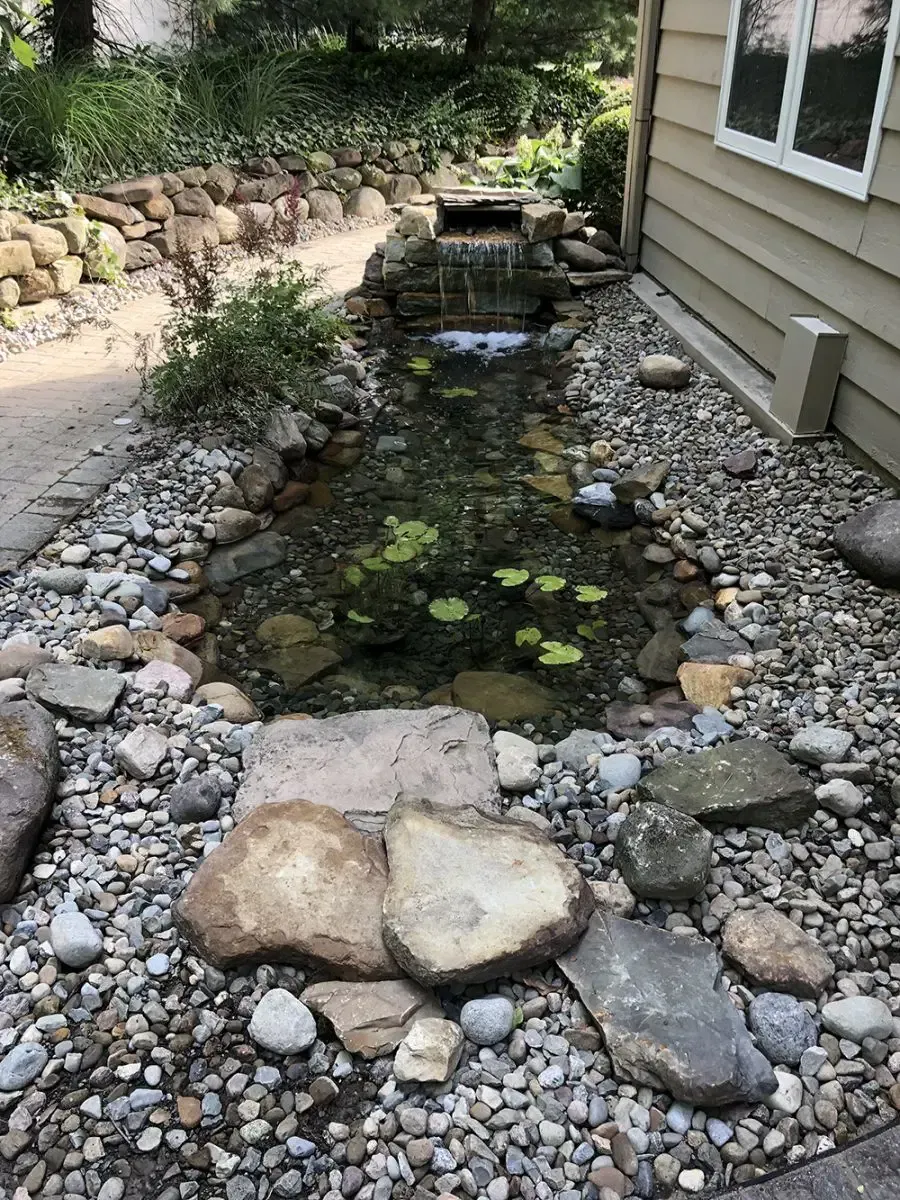 Narrow backyard pond with rocks, stepping stones, and a small waterfall beside a house.