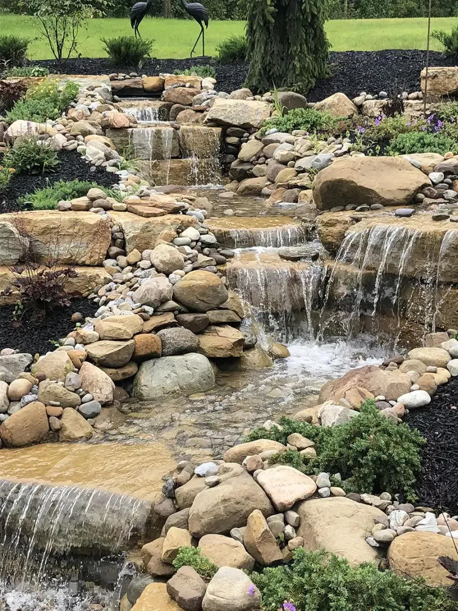 Tiered rock waterfall cascading into a landscaped pond with green grass and plants nearby.