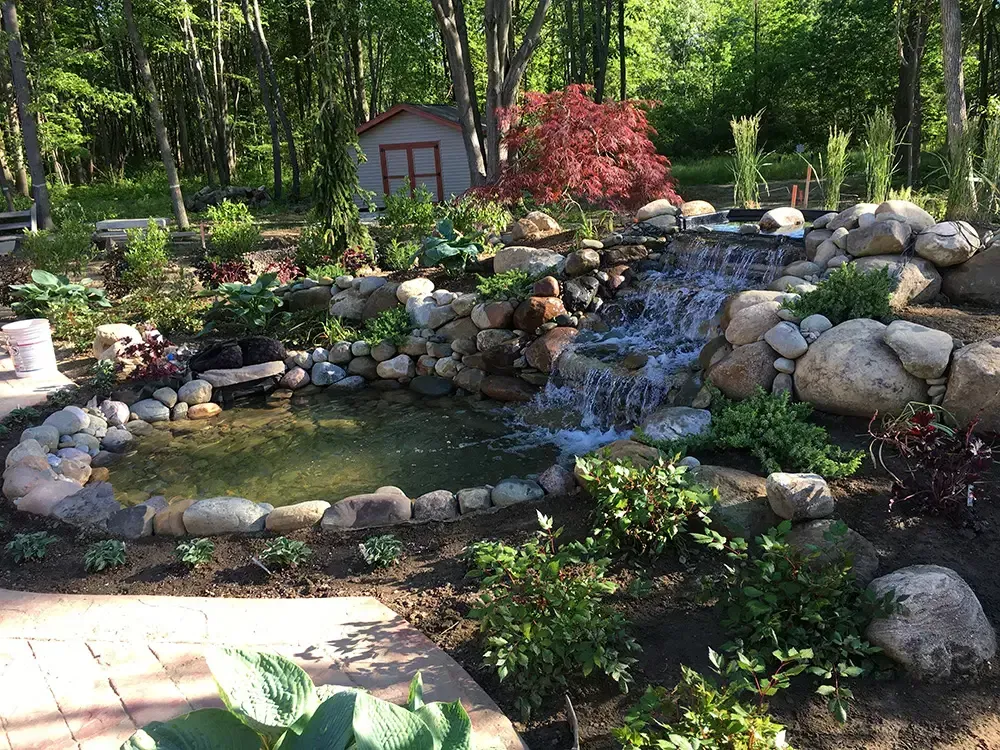 Stone garden pond with small waterfall, surrounded by flowers, rocks, and trees.