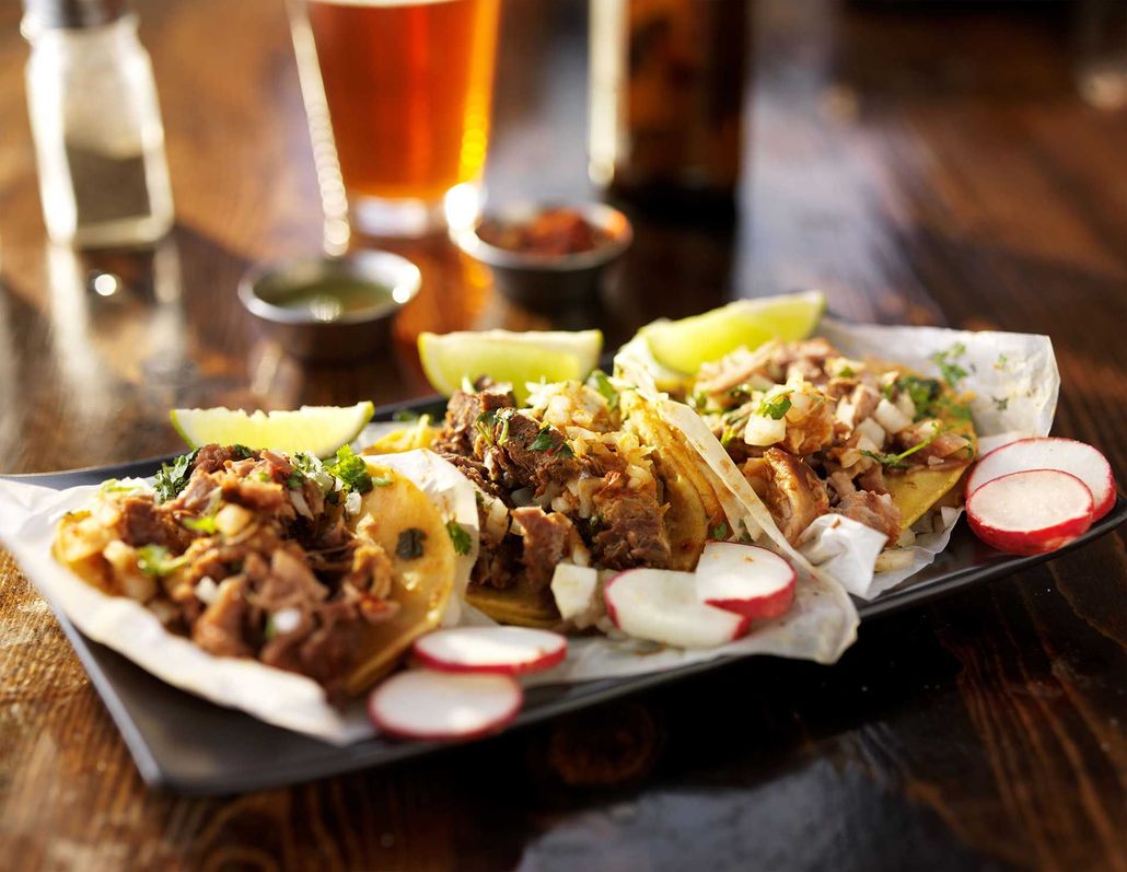 Three tacos with meat, cilantro, and onion on a plate with lime wedges and sliced radishes on a dark wood table.