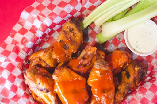 Buffalo wings in red sauce with celery and ranch dressing in a red and white checkered basket.