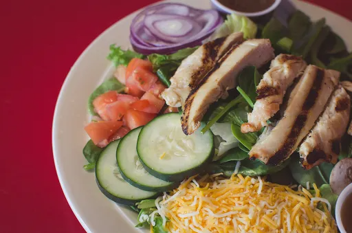 Salad with grilled chicken, tomatoes, cucumbers, red onion, and shredded cheese on a white plate.