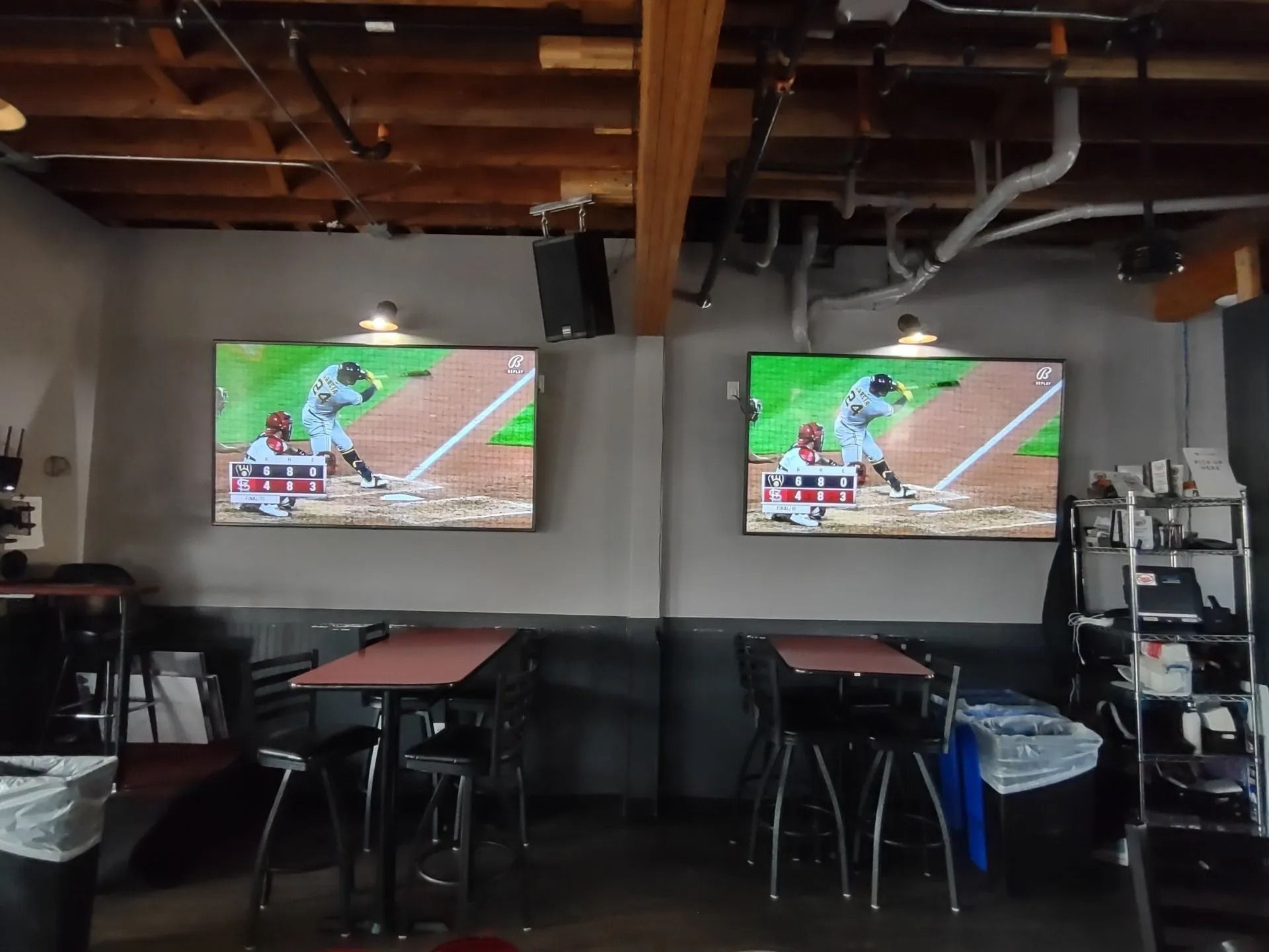 Two TVs showing a baseball game above tables in a bar. Gray walls, wood ceiling.