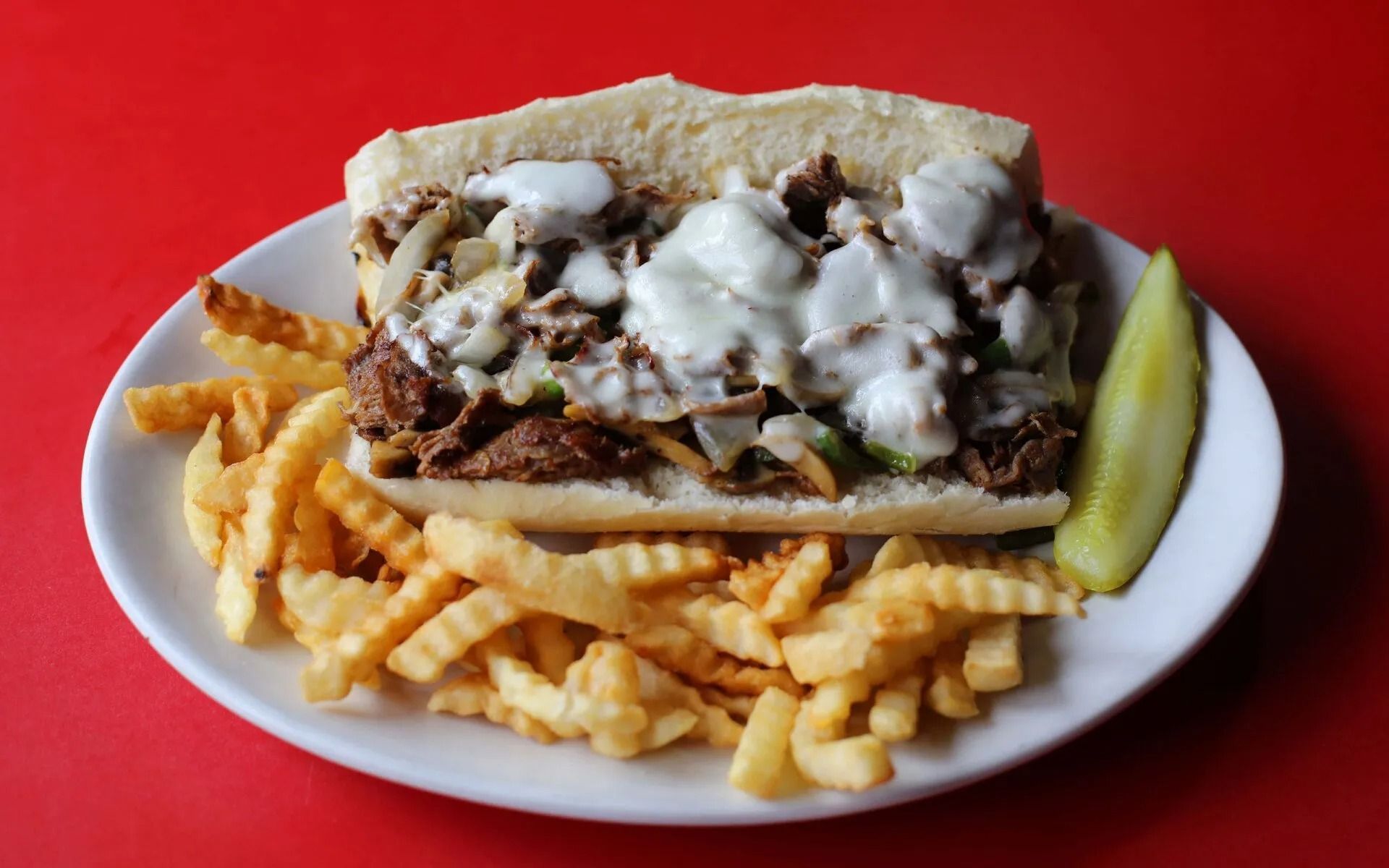 Cheesesteak sandwich with fries and a pickle on a white plate, red background.