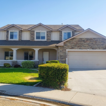 A two-story tan suburban house with stone veneer accents, a covered front porch, a two-car garage, and a manicured lawn.