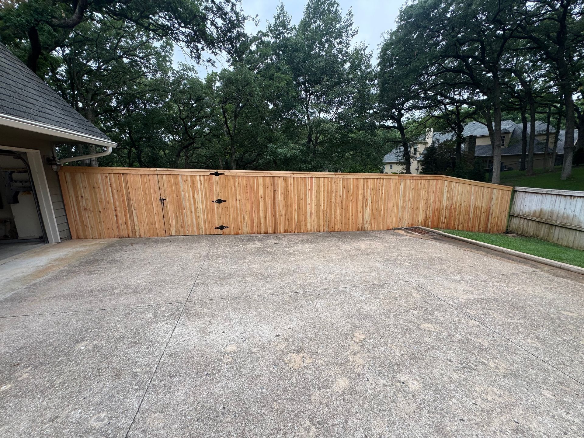 Wide gravel driveway beside a wooden privacy fence and a house garage, with trees in the background.