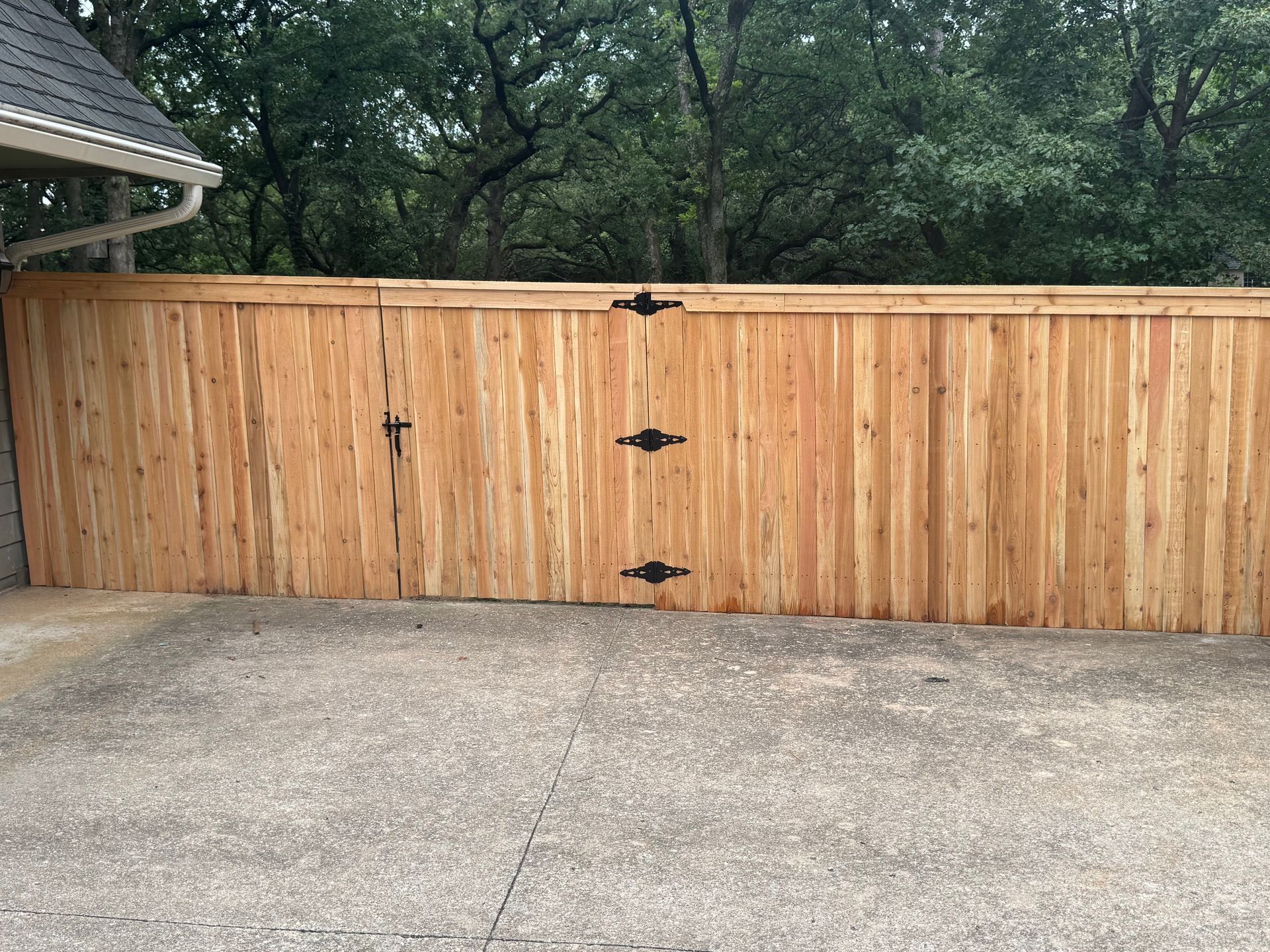 Wooden privacy fence with black hinges across a concrete driveway, backed by trees and part of a house roof.