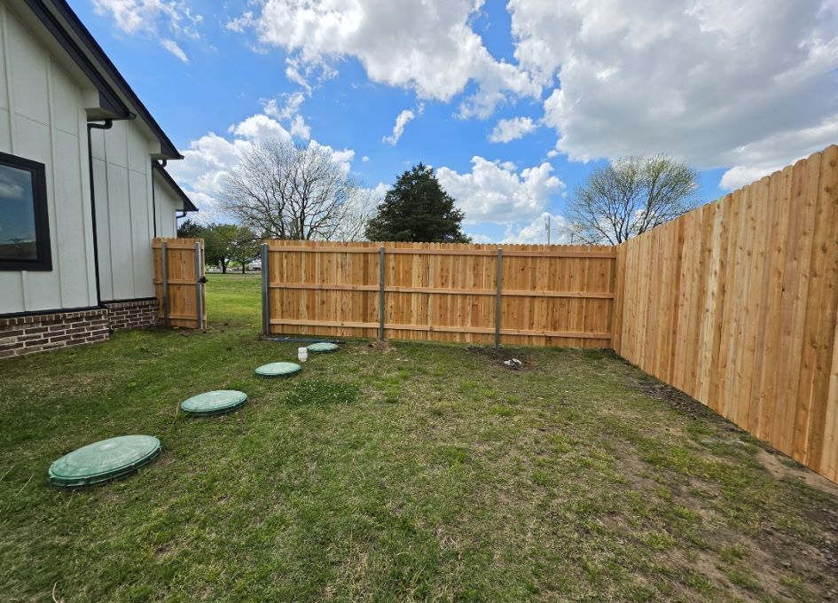 Small backyard with wooden fence, grass, stepping stones, and a house exterior under a cloudy blue sky