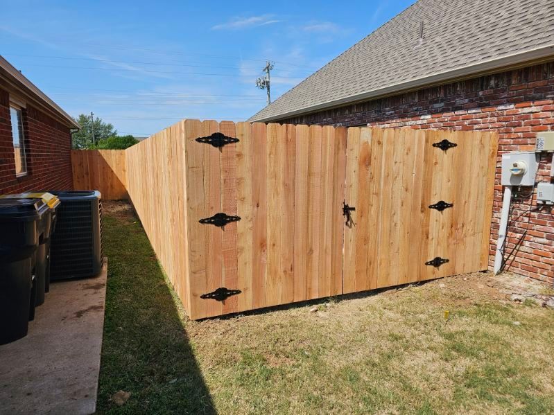 Wooden double gate with black hinges between a brick house and fence in a grassy yard