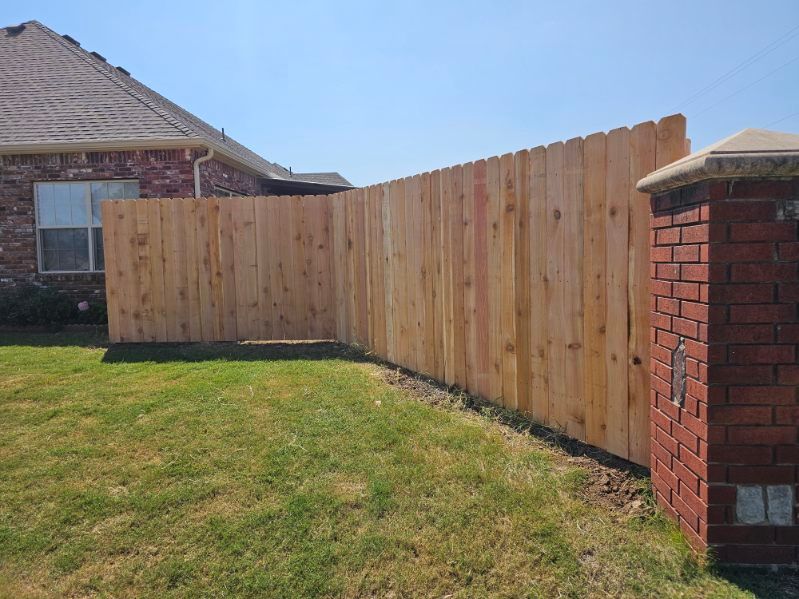 Wooden privacy fence along a grassy yard beside a brick post and house under a clear sky